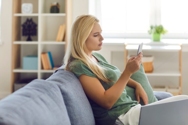 Side view of woman sitting on sofa at home, using mobile application on cellphone, chatting on social media, reading texts, shopping in online store. Modern gadgets or compulsive phone use concept